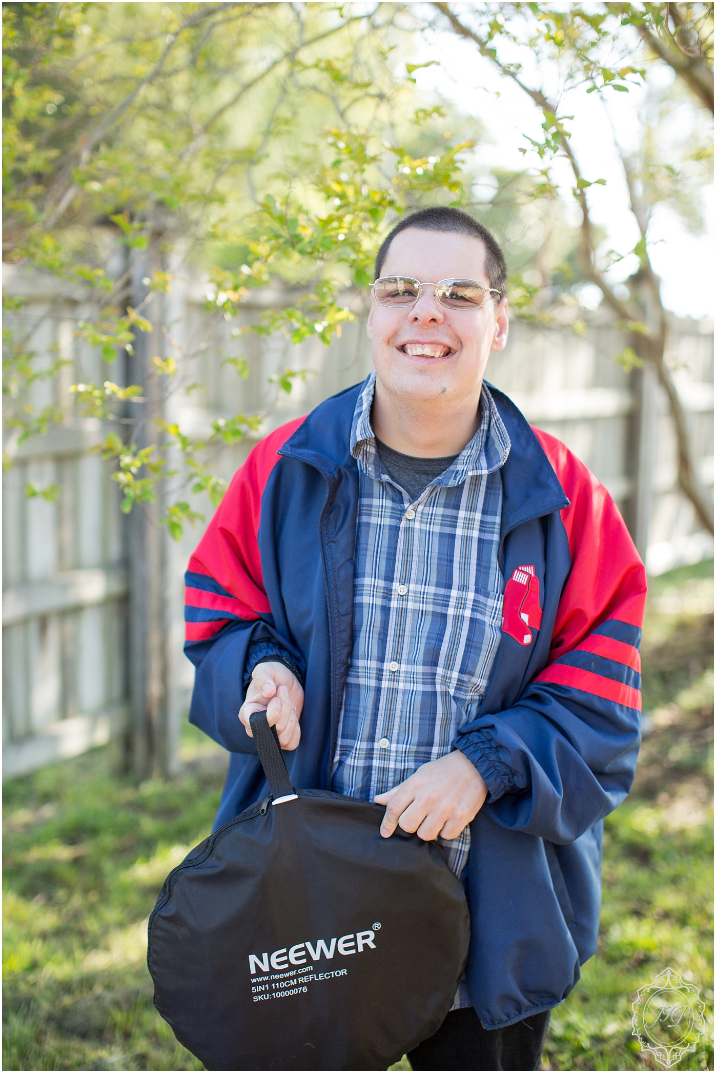 Autistic adult male wearing a striped shirt smiles at the camera while holding a reflector while standing in front of a fence and naturally lit tree in Columbia, South Carolina.
